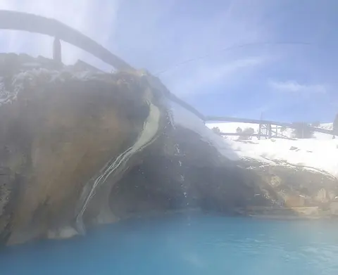 A hot spring with blue water and mineral deposits below a snowy bank.