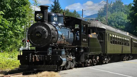 A historic steam train runs alongside a road with green trees in the background.