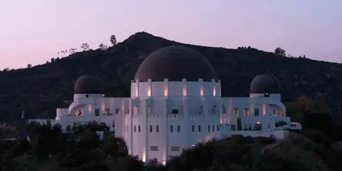 A large white observatory building with multiple domes sits in front of a dark hill at twilight.