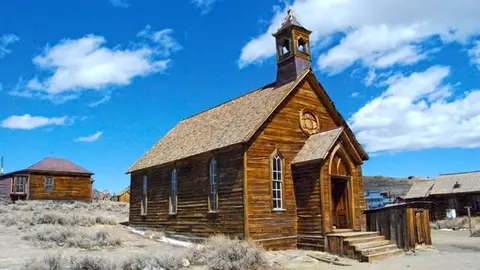 An old wooden church building stands against a bright blue sky with clouds.