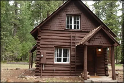 A brown log cabin stands among tall green trees.
