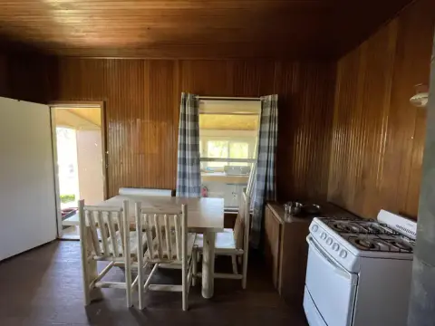 An interior shot of a rustic room with wooden paneling, a dining table, chairs, and a stove.