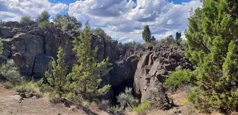 A rocky opening or fissure in the ground surrounded by green trees and bushes under a partly cloudy sky.