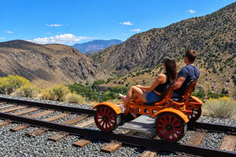 A couple rides a rail bike on railroad tracks overlooking a mountain canyon.