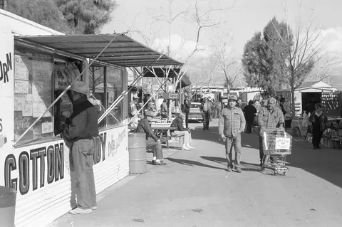 A black and white photograph of people walking near a food stand at an outdoor market.