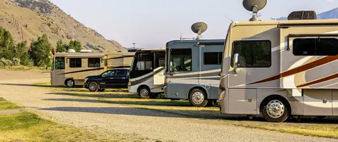 A row of recreational vehicles parked in a gravel lot with mountains in the background.