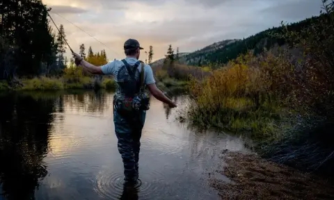 A person fly fishes in a river surrounded by trees and hills.