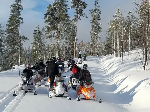 A group of people riding snowmobiles on a snowy trail through a forest.