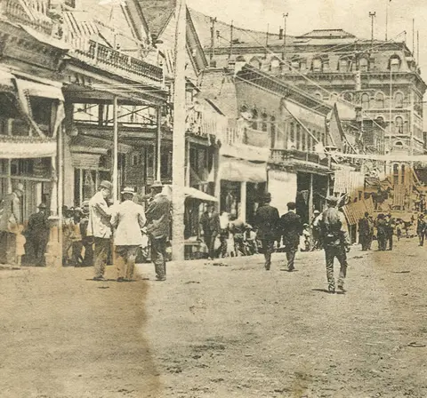 Historic black and white photo of people walking along a street with old buildings.