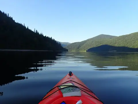 A view from the front of an orange kayak on a calm body of water surrounded by forested mountains.