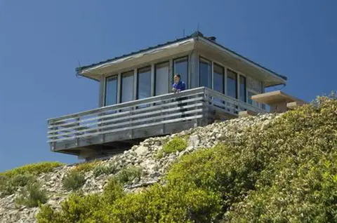 A person stands on the balcony of a small lookout building on a rocky hill.