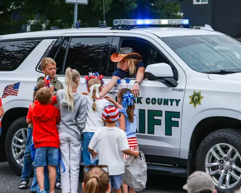 A group of children gather around the window of a sheriff's vehicle where an officer wearing a patriotic hat greets them.