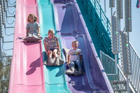 Three children smile as they slide down a colorful amusement park slide.