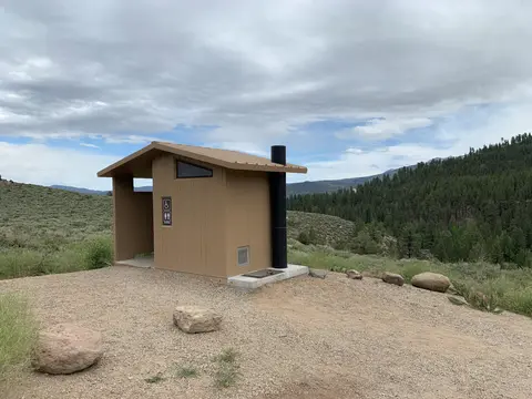 An outdoor toilet building sits in a mountainous, forested landscape under a cloudy sky.