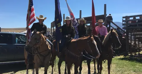 Four riders on horseback hold flags outdoors near wooden structures.