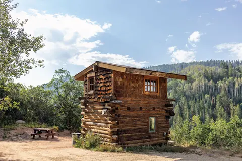 A wooden cabin stands in a forested mountainous landscape with a picnic table.