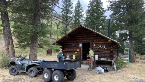 Two people are working outside a rustic log cabin in a wooded area with an ATV and utility trailer.
