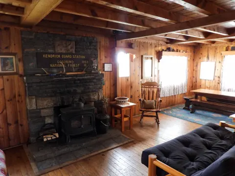 An interior view of a rustic wooden cabin with a stone fireplace and wood-burning stove.