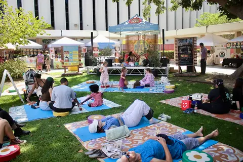 People relax and picnic on mats on a grassy area in front of an outdoor stage.