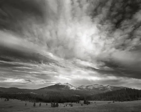 Black and white landscape photo showing mountains, forests, and a dramatic cloudy sky.
