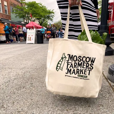 A person holds a canvas tote bag filled with greens at an outdoor market.