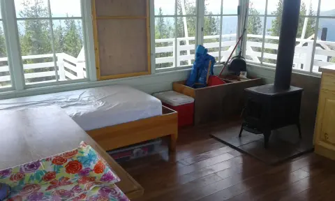 An interior view of a cabin featuring a bed, wood-burning stove, and large windows.