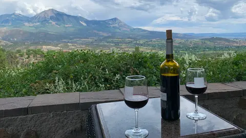 A bottle of wine and two glasses sit on an outdoor table overlooking a mountain landscape.