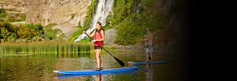 Two people paddle board on a river with a waterfall in the background.