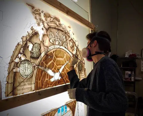 An artist wearing a respirator works on a detailed stained glass window panel.