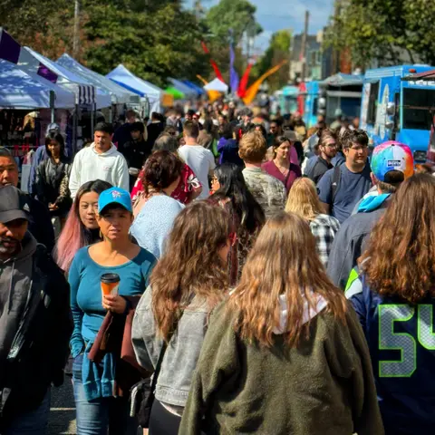 A large crowd of people walks along an outdoor street market with tents and food trucks.