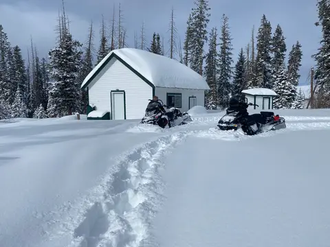 A snowy cabin surrounded by snowmobiles and evergreen trees.