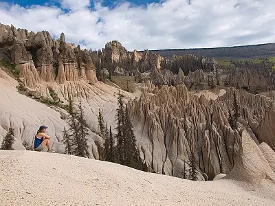 A person sits overlooking a landscape of eroded rock formations and sparse trees under a cloudy sky.