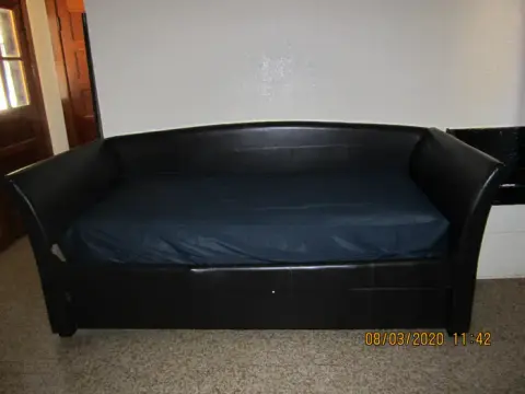 A black daybed with a dark mattress sits against a white wall indoors.