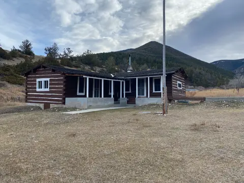 A log cabin sits in a grassy field with mountains in the background.