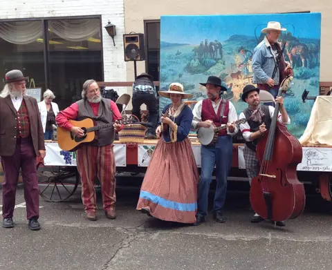 A group of musicians in historical costumes play instruments on a stage outdoors.
