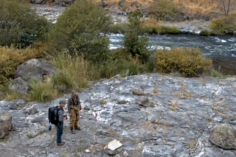 Two anglers stand on rocky ground near a flowing river.