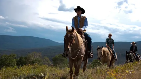 People ride horses across a grassy hillside with mountains in the background.