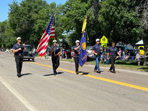 A color photograph shows a group of five people marching down a paved street carrying flags and rifles during a parade.