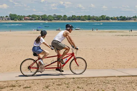 A couple rides a tandem bicycle along a path near a beach and water.