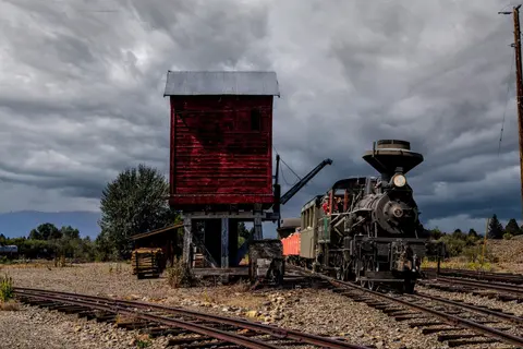 A steam locomotive parked next to a red water tower on a cloudy day.