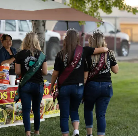 Three young women wearing tiaras and sashes walk away from a food tent.