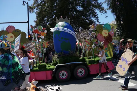A decorated parade float featuring a globe and flowers moves down a street with people walking alongside.