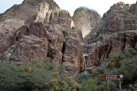 A waterfall flows down between rocky mountain peaks with a trail sign in the foreground.