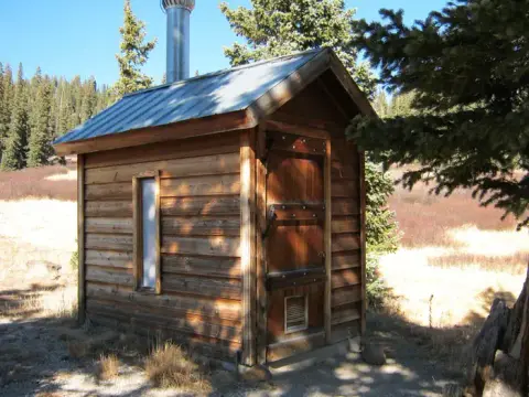 A small wooden outhouse stands in a mountainous landscape.
