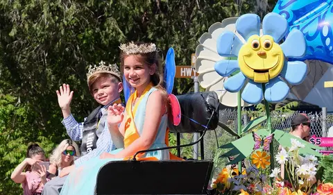 Two children wearing crowns and sashes wave from a parade float decorated with flowers.