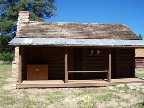 A historic log cabin with a stone chimney and a covered porch stands in a grassy area.