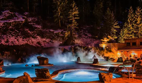 A night view of outdoor hot spring pools surrounded by snow-covered rocks and trees.