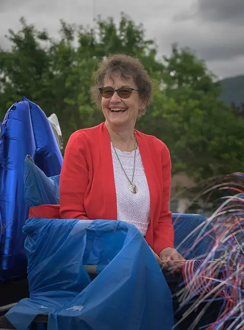 A smiling woman wearing sunglasses sits outdoors near blue decorations.