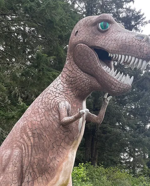 A large dinosaur statue stands outdoors in front of a forest.