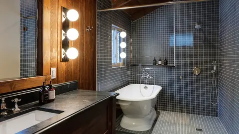 A modern rustic bathroom with a white freestanding tub, dark tiled walls, and a wooden vanity.
